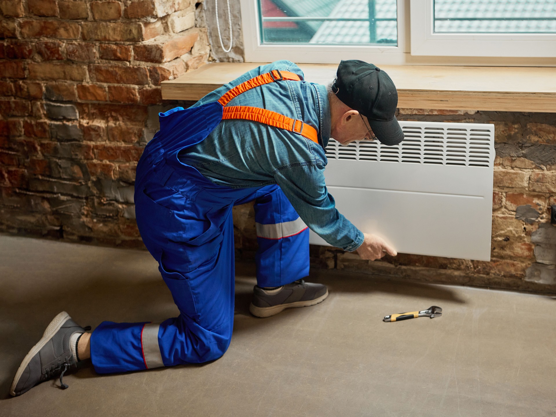 Technician installing white electric convector heater on brick wall indoors